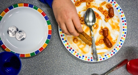 A person is eating from a plate of half-eaten toast and beans, with another plate with two silver coins on next to it 