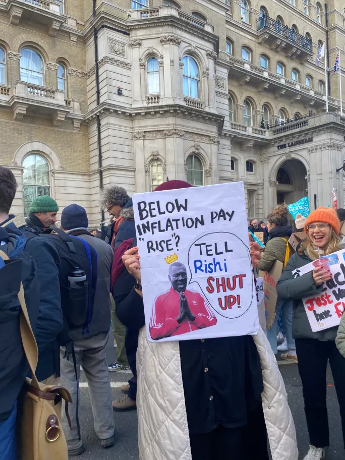 Gina holds a placard with a picture of Stormzy reading "Below inflation ay rise? Tell Rishi shut up"