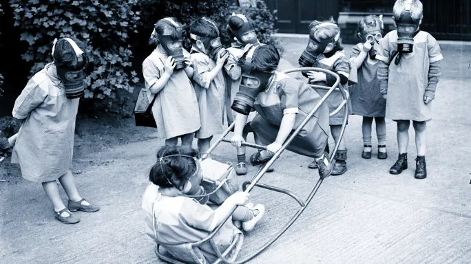 Children in playground, wearing gas masks. Two play on seesaw.