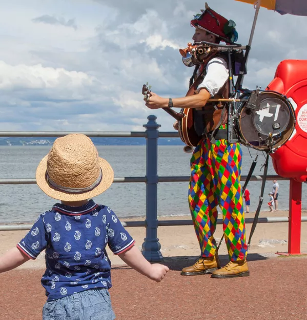 Solo Baby Approaches One-man Band On A Seafront Promenade - Independent Learners