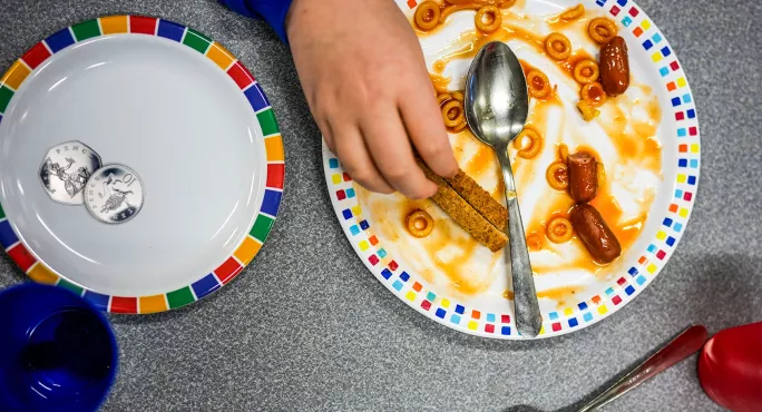 A person is eating from a plate of half-eaten toast and beans, with another plate with two silver coins on next to it 