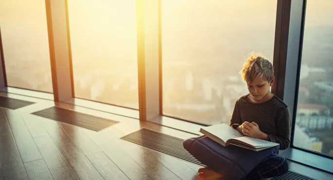 Boy Reads Book In Front Of Large Window In Luxury Flat