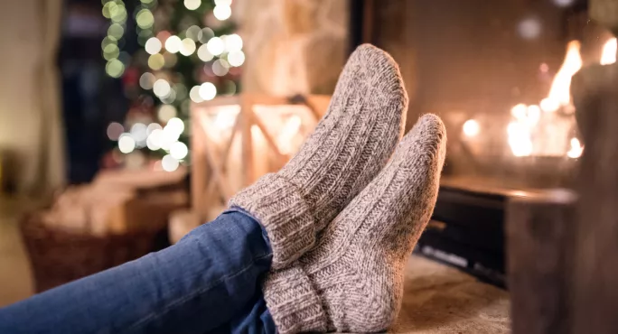 A Woman, Wearing Thick Socks, Puts Her Feet Up By The Fire At Christmas