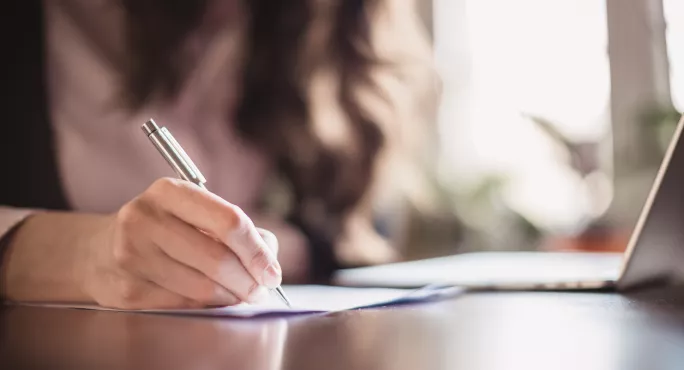 A Woman, Writing With Pen At A Desk, With A Laptop Next To Her